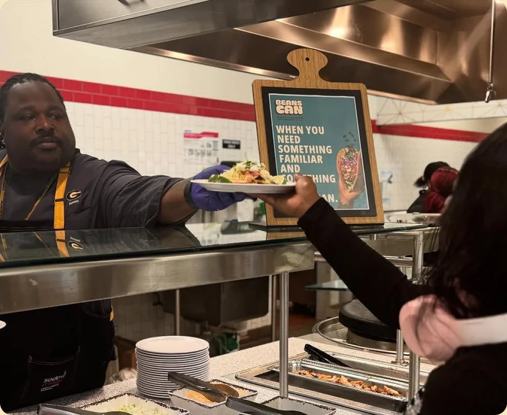 Staff member serving a bean dish next to a Beans Can promotional sign